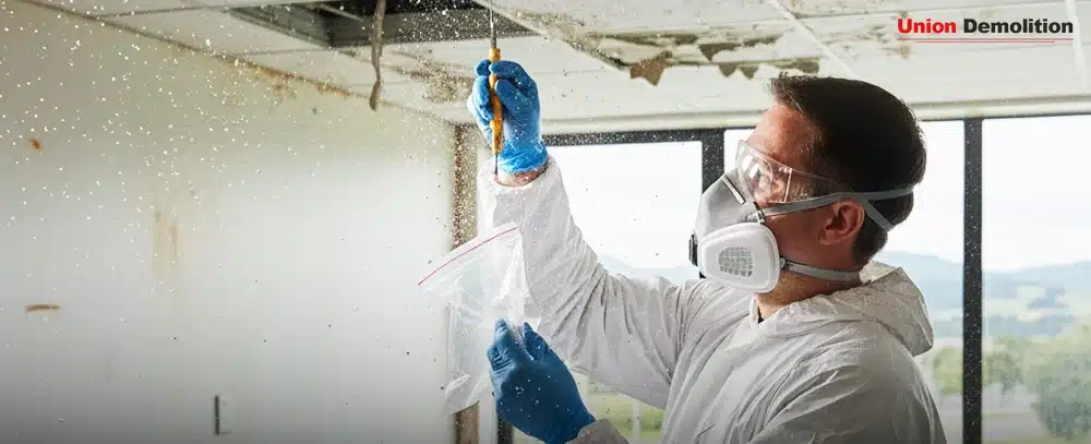 Worker in PPE collects a sample from a deteriorating ceiling, wearing a respirator, goggles, and gloves for safety.