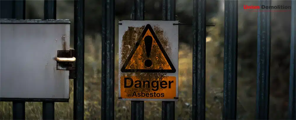A weathered yellow warning sign on a black metal fence that reads "Danger Asbestos" with a large exclamation point symbol. The background is blurred with natural greenery.